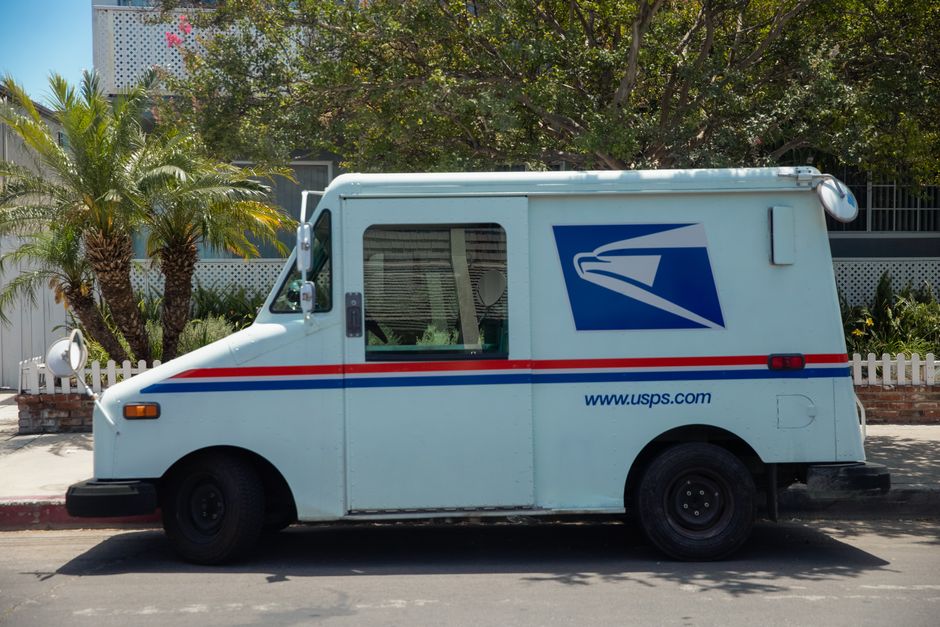 A white USPS mail delivery van parked on a street in daytime with a backdrop of green trees and a white picket fence. The van features the blue USPS eagle logo, red and blue stripes along its side, an