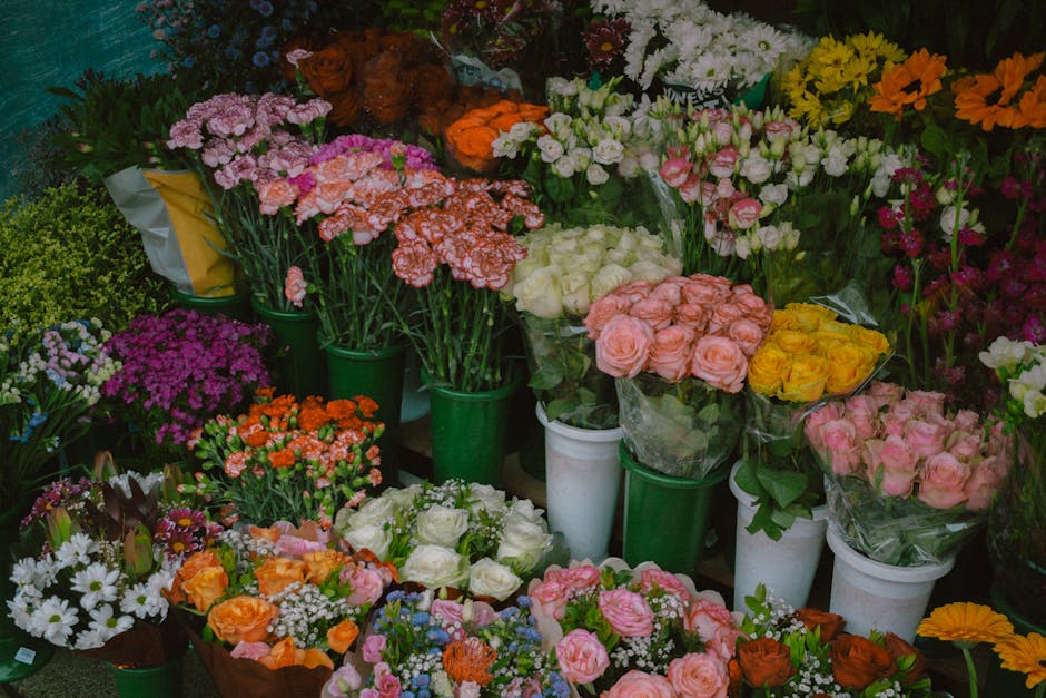 A vibrant display of fresh flowers arranged in white and green vases and wrapping, showcasing a variety of blooms including pink, white, yellow, orange, and purple flowers. The bouquet arrangements fe
