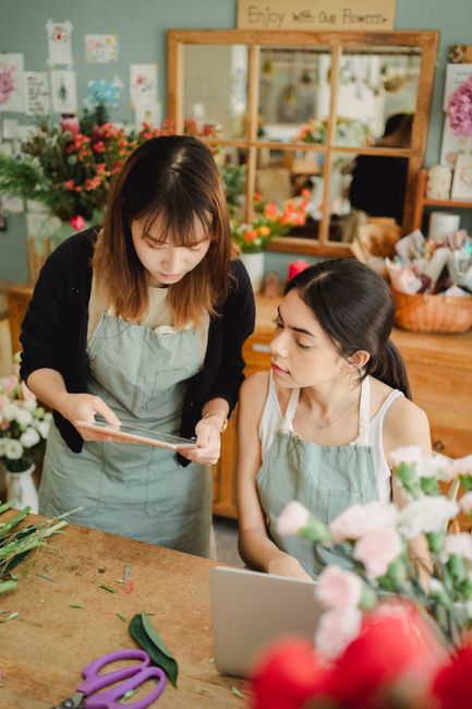 Two women working in a flower shop, with one standing and holding a tablet and the other seated at a wooden worktable covered with a variety of fresh flowers. The arrangement includes pastel pink rose