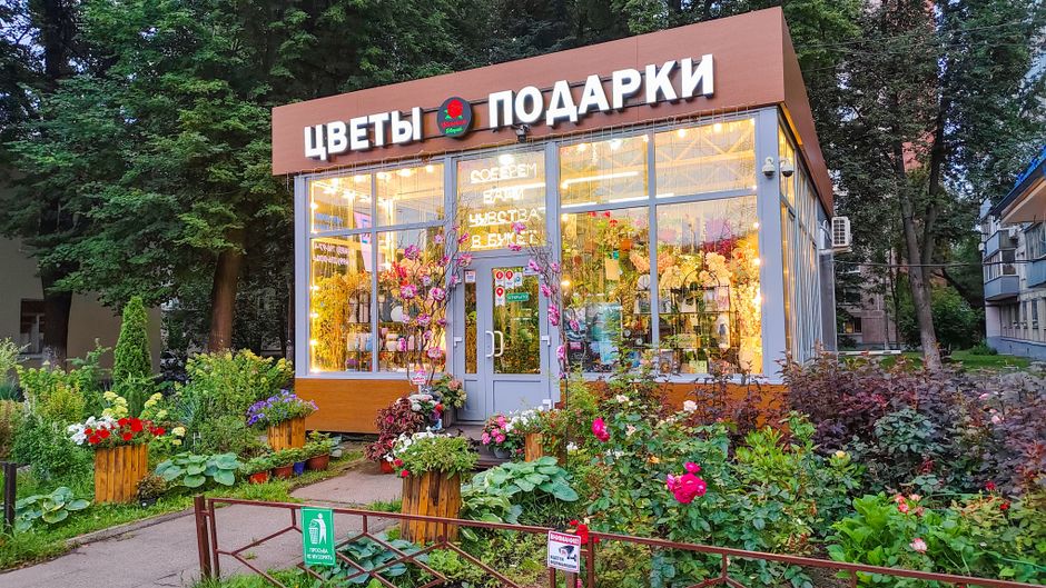 The image features the exterior of a flower shop located on Station Road in Highams Park, with large glass windows and a sign in Cyrillic script. The shop is adorned with a variety of potted plants an