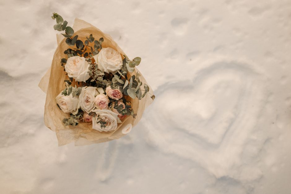 A bouquet of fresh white and pale pink roses, accented with green eucalyptus leaves, wrapped in light brown kraft paper, resting on snow with a heart shape drawn in the snow nearby. The arrangement is