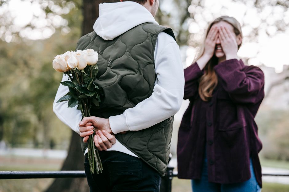 A person holding a bouquet of pale peach and cream roses behind their back, with green stems and leaves visible, while another person in a purple jacket covers their face with their hands in a park se