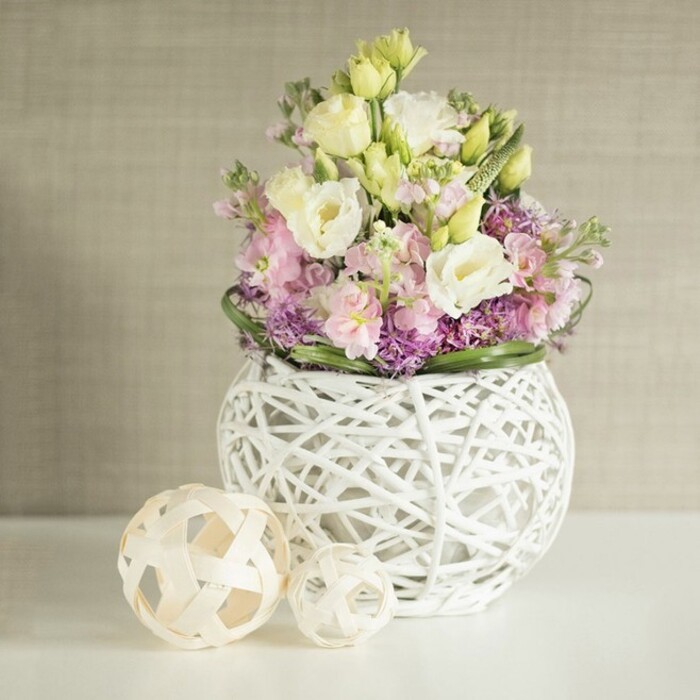 Flooded with diffused natural light, a lavish floral centrepiece crafted by local Highams Park florists brings the promise of spring indoors. The abundant bouquet emerges from a distinctive spherical vase, its crisp white openwork-fashioned from woven rattan-speaking of modern openness and organic craft much like the marketplace finds near The Avenue. Within, ruffled, creamy white and buttery yellow lisianthus mingle closely with tall, pastel pink stocks, giving the design elevation and gentle flair. The lower portion is completed by a cushion of rich purple fillers-resembling asters or dainty alliums-arranged thoughtfully to add captivating texture and balance, all set within a frame of glossy deep green leaves that recall the trees of Larks Wood. To one side, two decorative spheres, meticulously crafted from pale rattan or wood in open, geometric lattice patterns, lend the scene additional warmth and handcrafted charm. Against a calm greige backdrop with a subtle, fabric-like texture, the bouquet's varied tones and intricate structures capture a sense of serene luxury and freshness. Poised for wedding breakfasts, elegant gatherings, or refined seasonal displays, this arrangement celebrates Highams Park's love for nature, artistry, and local design inspiration.